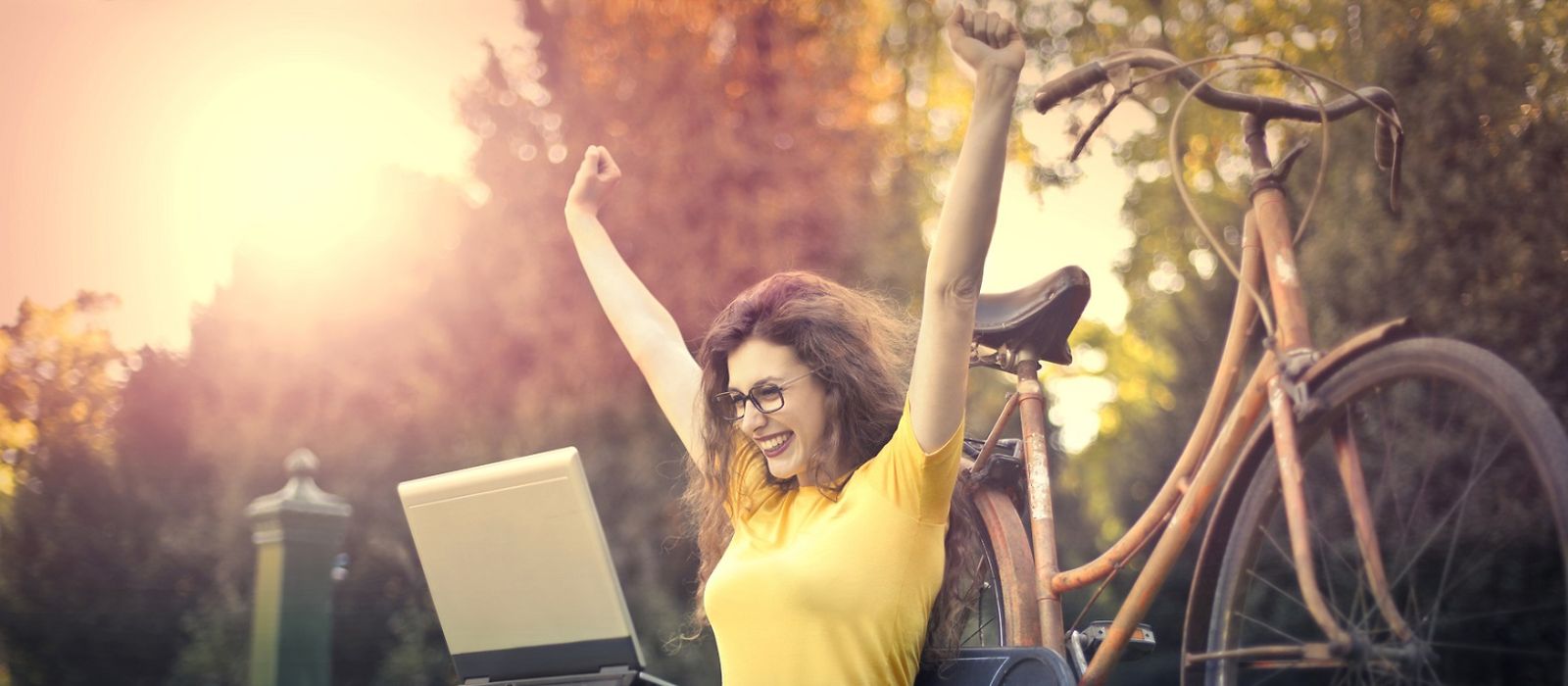 Frau sitzt auf einer Wiese, freut sich, Arme hochgerissen, Laptop auf dem Schoss, Fahrrad im Hintergrund