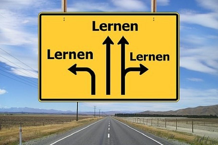 Landstraße, Felder und Berge, blauer Himmel, leichte Wolken, gelbes Schild mit Pfeilen und den Worten Lernen, Lernen, Lernen 