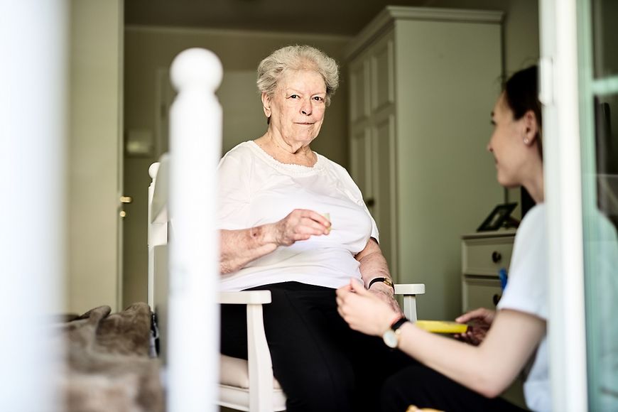 Seniorin im Zimmer, sitzend auf einem Stuhl, eine jüngere Frau gegnübersitzend