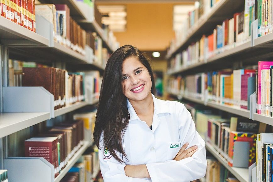 Frau in einer Bibliothek zwischen zwei Bücherregalen, die Frau lächelt, Arme sind vor der Brust verschränkt