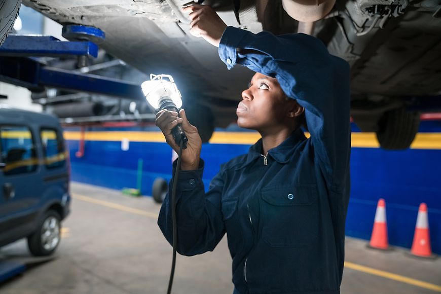 Eine weibliche Mechatronikerin arbeitet in der Autowerkstatt unter einem Auto