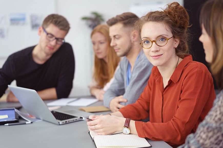 Eine Frau im Arbeitskontext schaut in die Kamera, mit ihr sitzen weitere Personen am Tisch die leicht verschommen sind