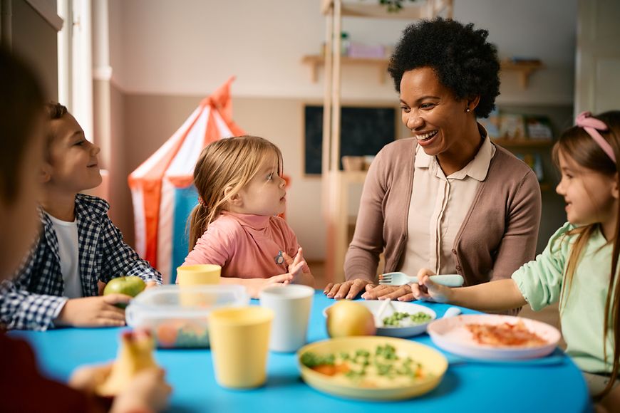 Frau sitzt mit Kindern am Mittagstisch und lacht. 