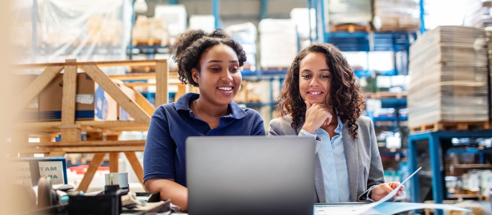 Zwei Frauen sitzen in einer Lagerhalle am Laptop.