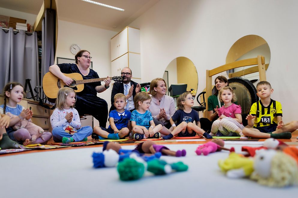 Lisa Paus sitzt mit Kindern auf dem Boden. Im Hintergrund spielt jemand Gitarre