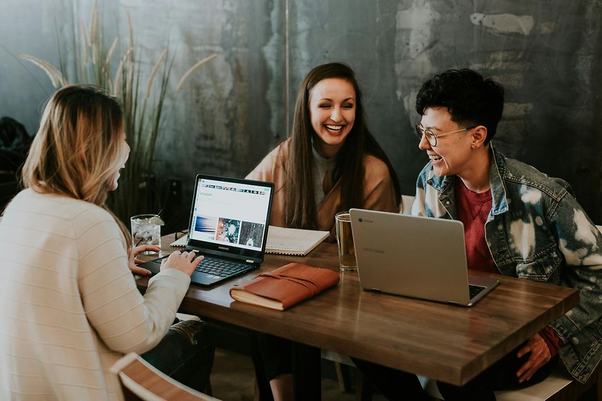 Drei Frauen sitzen mit Laptop am Tisch und lachen